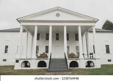 A Large White Plantation House With Columns And Pumpkins And Rocking Chairs On The Front Porch.