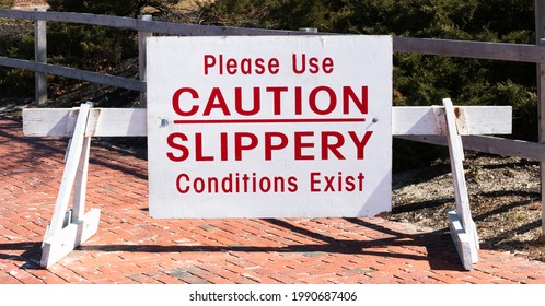 A Large White Sign On A Wood Barrier With Red Text Reads Please Use Caution Slippery Conditions Exist At The Entrance Of A Park On A Sunny Day.