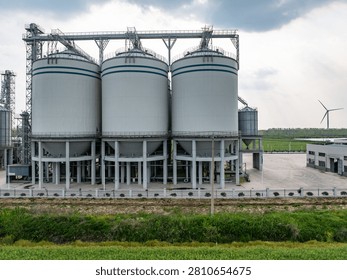 Large white silo complex with wind turbine and green fields under cloudy sky