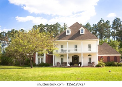 Large White Southern Style Coastal Home With A Front Porch. The Yard Features A Grass Lawn And Large Oak Trees.