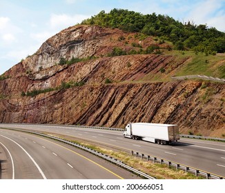A large white trailer truck drives through a mountain pass with steep sedimentary rock to the sides of the road.