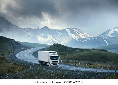 Large white transport truck transporting commercial cargo in semi trailer running on turning way highway road with scenic mountains mountaineous scenery in background.