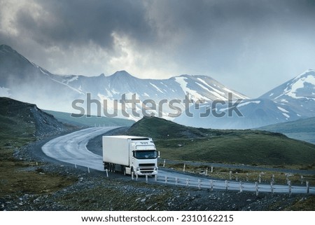 Large white transport truck transporting commercial cargo in semi trailer running on turning way highway road with scenic mountains mountaineous scenery in background.