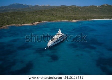A large white yacht is anchored off the coast of Sardinia in the background. Coastline, green mountains, blue sky. White Mega yacht moored off the coast of Sardinia on blue water.