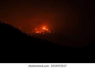 A large wildfire burns intensely on a forested mountain at night, casting a bright, orange glow that illuminates the smoke and surrounding dark hillsides. The fire appears expansive and menacing again - Powered by Shutterstock