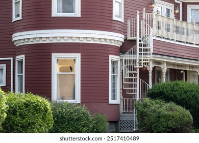 A large wooden building with burgundy clapboard and white trim. The wall is round with three windows. There's a white metal spiral staircase and railing with a balcony over the facade of the house.  - Powered by Shutterstock