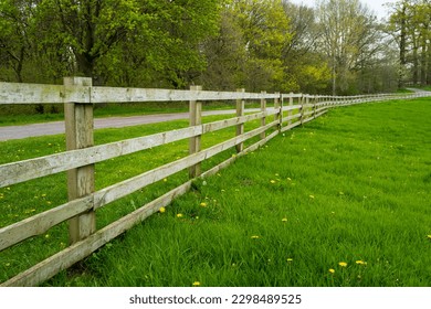 Large wooden fence seen at the perimeter for a large meadow with a public path seen following the fence into a forest area.