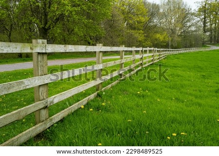 Large wooden fence seen at the perimeter for a large meadow with a public path seen following the fence into a forest area.