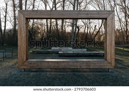 Large wooden rectangular frame structure in a quiet park with benches, bare trees, grass, and soft evening sunlight.