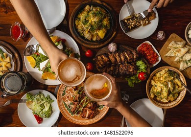 A Large Wooden Table Generously Covered With Delicious National Dishes, With Friends Sitting And Drinking Light Beer From Glasses