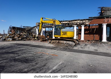 A Large Yellow Backhoe In The Parking Lot In Front Of A Building Being Demolished After Being Destroyed In A Fire