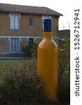 Large yellow bottle positioned in a town square opposite a yellow mustard coloured painted house, overcast day white skies.