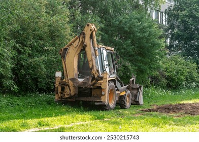 Um grande veículo de construção amarelo e laranja está estacionado em um campo gramado. O veículo é uma retroescavadeira, e é cercado por árvores. A cena é pacífica e serena