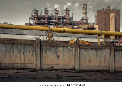 Large Yellow Pipe On Top Of A Large Concrete Wall In Front Of Giant Smokestacks Of An Abandoned Industrial Blue Collar Factory In A Small Rust Belt Town In West Virginia