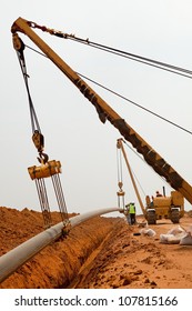 Large yellow side boom pipe layer or stringer industrial machine on orange dusty sandy outdoor construction site laying a pipe in a trench watching by workers