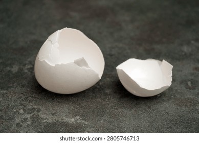 A larger broken eggshell rests next to a smaller piece. The shells are white and sit on a gray stone surface, hinting at recent activity in a kitchen or cooking area.