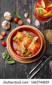 Lasagna Rolls In Clay Bowl With Tomato Sauce And Arugula On Aged Wooden Table With Basil Leaves, Cherry Tomatoes, Garlic And Red Cocktail. Overhead View