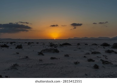 The last rays of the sun dip below the horizon, casting a vibrant golden glow behind dark mountain ranges. Somber clouds and dark sandy terrain dominate the frame, creating a moody atmosphere of endings and quiet reflection.