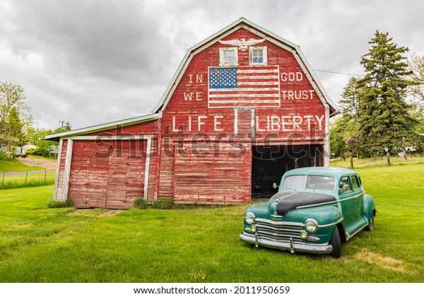 Latah, Washington,\
USA. May 24, 2021. Vintage Plymouth Super De Luxe automobile and a\
weathered red a barn.