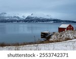 Late autumn country, covered by snow. Water of Norweigan sea (Arctic ocean), reflecting cloudy blue sky. Red house on the coast. Harstad, Norway.