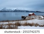 Late autumn country, covered by snow. Water of Norweigan sea (Arctic ocean), reflecting cloudy blue sky. Red house on the coast. Harstad, Norway.