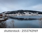 Late autumn country, covered by snow. Water of Norweigan sea (Arctic ocean), reflecting cloudy blue sky. Houses on the coast. Harstad, Norway.