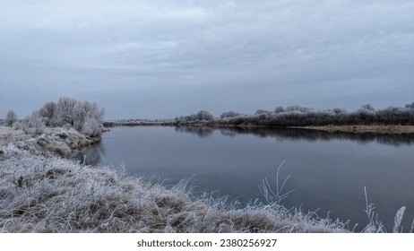 In late autumn, on grassy banks of the river, trees stand without leaves. After a night frost, grass, bushes and tree branches are covered with frost. The trees are reflected in the calm water. Cold - Powered by Shutterstock