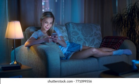 Late At Night Beautiful Woman Lying On Her Sofa In The Living Room, Using Her Smartphone And Smiling.