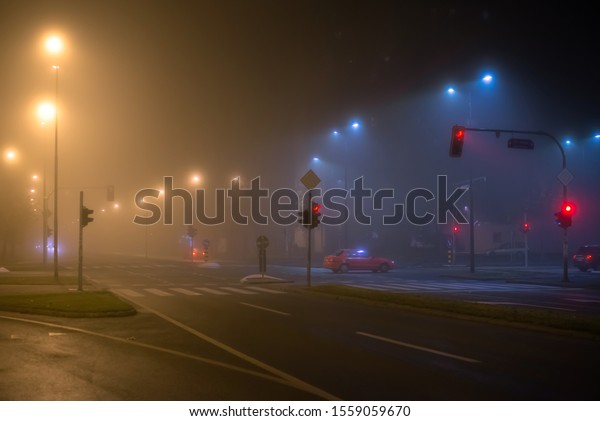 Late night in the city\
streets with fog, red stop lights and a red car. Reduced\
visibility, bad weather. 