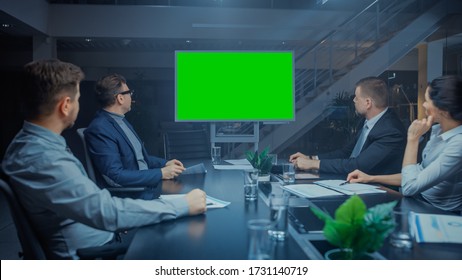 Late At Night In Corporate Meeting Room: Board Of Directors, Executives And Businesspeople Sitting At Negotiations Table, Talking And Using Green Mock-up Screen Wall TV For Video Conference Call.