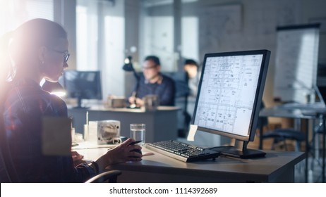 Late at Night/Early Morning in the Engineering Bureau Female Designer Works on a Personal Computer that Shows Blueprints of Her Project. In the Background Her Colleagues Working. - Shutterstock ID 1114392689