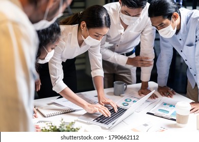 Late Working Of Asian Teamwork Business People Working In Office With New Normal Lifestyle. Group Of Man And Woman Wear Protective Face Mask To Prevent Covid Infection, Meeting On Table At Night.