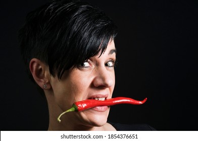 Lateral Head Portrait Of A Young Black-haired Woman In Front Of A Black Background Holding A Long Red Chilli Pod Across Her Teeth And Looking Skeptically Into The Camera.