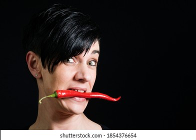 Lateral Head Portrait Of A Young Black-haired Woman In Front Of A Black Background Holding A Long Red Chilli Pod Across Her Teeth And Looking Skeptically Into The Camera.