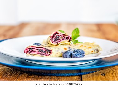 Lateral View Of Crepe With Blueberry Jam Filling Cut In Half And Decorated With Mint Leaves And Frozen Blueberries On White And Blue Plates.