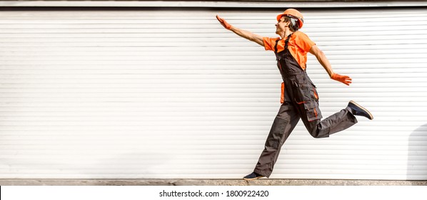 Lateral view of young happy laughing caucasian man builder construction worker in a safety helmet are jumping in front of the roller door lifting gates. Copy space for you text and logo - Shutterstock ID 1800922420
