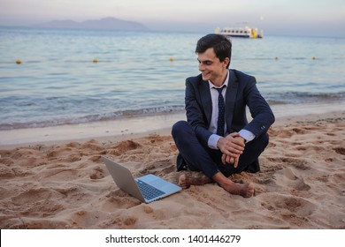 Lateral View Of A Young Man In Suit With Laptop Sitting On The Beach Working On The Seaside