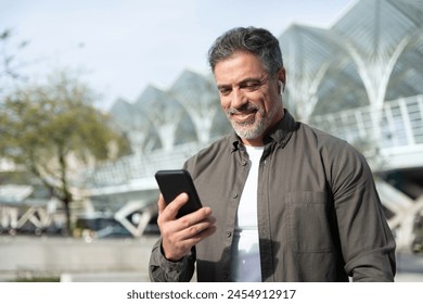 Latin 40s smiling mature hispanic business man using smartphone cellphone app, earphones for voice call app. Middle age entrepreneur businessman holding mobile cell phone, watching, listening outdoors - Powered by Shutterstock