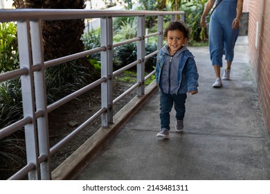 Latin American Boy Walks Alone In Front Of His Mother. He Smiles At The Camera. Concept Going For A Walk, Motherhood