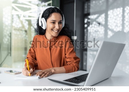 Latin american business woman with curly hair and headphones watching online training course at workplace, woman writing information happy and satisfied with the results of professional development.