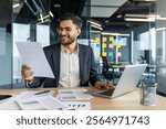 Latin American businessman reviews important documents while working on his laptop in a modern yet professional office environment. This portrays professionalism, productivity, and a positive work