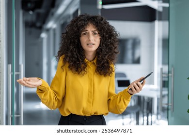 Latin American businesswoman in office corridor holding phone, wearing yellow blouse, showing confusion or frustration. Raised hands depict communication challenge or misunderstanding. - Powered by Shutterstock