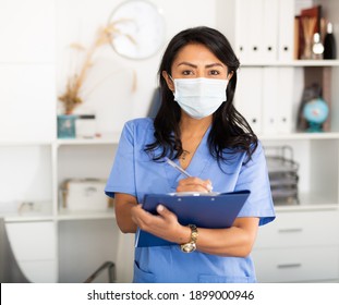 Latin American Female Nurse Wearing Face Mask While Writing Notes And Reading Something On Clipboard At Her Office During Coronavirus Quarantine