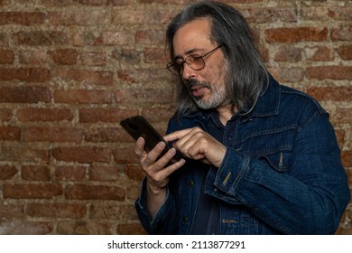 Latin American Man (44) With Long Gray Hair Checks Messages On His Cell Phone. Concept Of Technology