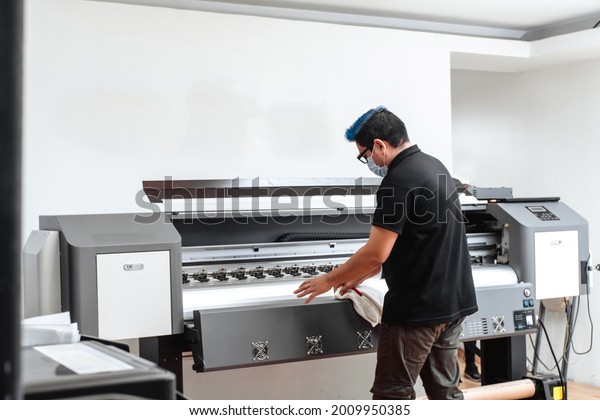 Latin American man with mask\
cleaning large format plotter. concept business and corona\
virus.