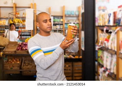 Latin American Man Reading Jar Contents On Label While Shopping In Food Department Of Supermarket