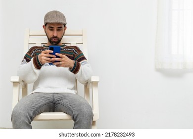 Latin American Man Sitting On A Chair In His House With His Cell Phone