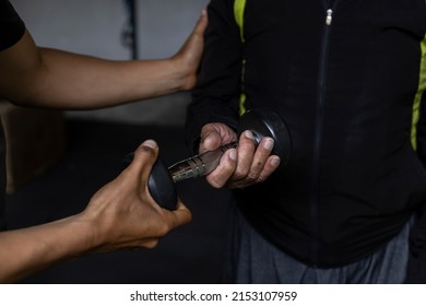 Latin American Senior Male With His Unrecognizable Personal Trainer Do Rehabilitation And Therapy Exercises Holding A Dumbbell In The Gym. Healthy Lifestyle Concept.