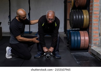 Latin American Senior Male With His Personal Trainer Do Rehabilitation And Therapy Exercises Holding A Kettlebell With Both Hands In The Gym. Healthy Lifestyle Concept.