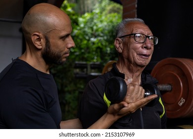 Latin American Senior Male With His Personal Trainer Do Rehabilitation And Therapy Exercises Holding A Dumbbell In The Gym. Healthy Lifestyle Concept.